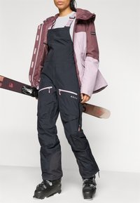 Conjunto de esqui com uma jaqueta impermeável bordeaux e rosa clara, calças de esqui pretas com bolsos com fecho éclair e botas de esqui pretas.