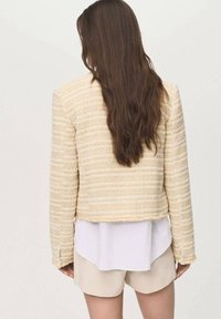 Yellow textured cropped jacket with long sleeves, woven detailing, and frayed hem, paired with a white shirt and beige shorts.