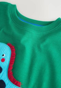 Jersey verde con cuello de canalé; presenta un diseño de aplicación de una criatura azul con acentos rojos y detalles faciales bordados.