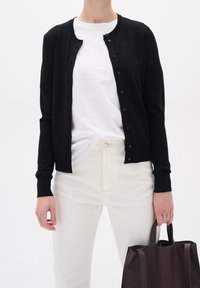 Person som bär en svart knappad cardigan över en vit t-shirt och krämfärgade byxor, håller en mörkbrun tote-väska vid sin sida.