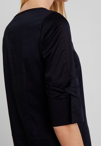Femme portant une chemise noire à manches longues, manches retroussées, vue de dos et de côté sur un fond clair.
