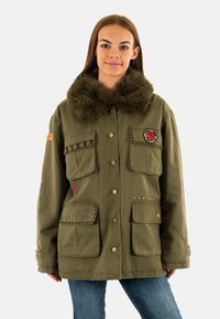 Giacca parka verde oliva, caratterizzata da un colletto in pelliccia, numerose tasche, cuciture decorative e toppe. Realizzata in tessuto resistente.