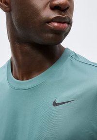 Κοντινό πλάνο στο κάτω μέρος του προσώπου και του λαιμού ενός άνδρα που φοράει ένα ανοιχτό τιρκουάζ μπλουζάκι Nike με μαύρο λογότυπο Swoosh στο στήθος.