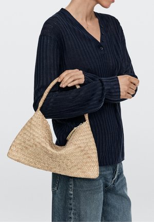 Osoba ubrana w granatowy sweter w prążki i dżinsy, trzymająca jasnobrązową plecioną torebkę z zakrzywioną rączką na ramieniu.