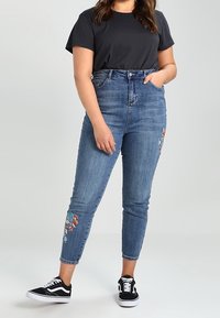 Džíny s vysokým pasem modré barvy mají na levé noze květinové vyšívání, doplněné černým tričkem a černými teniskami.