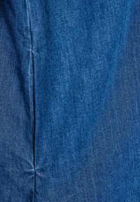 Close-up van blauw denimstof met zichtbare diagonale weefstructuur en een verticale naad aan de linkerkant.