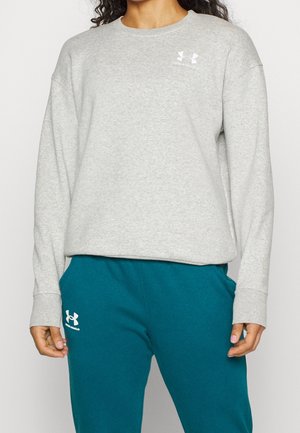 Nainen, jolla on päällään vaaleanharmaa Under Armour -collegepaita ja sinivihreät Under Armour -collegehousut, seisomassa kädet rennosti sivuilla.