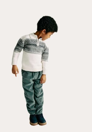 Jonge jongen die naar links onder kijkt, gekleed in een witte trui met patroon, groene ribfluwelen broek en zwarte laarzen, tegen een lichte achtergrond.