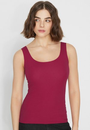 Femme aux cheveux bruns courts et ondulés, portant un haut sans manches rose foncé, regardant légèrement vers sa gauche sur un fond clair uni.