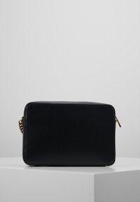 Clutch bag rettangolare nera in pelle con tracolla dorata e zip dorata, posata su un piedistallo bianco contro uno sfondo grigio.
