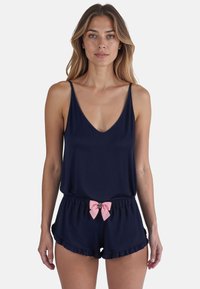 Mono de marina con escote en V, tirantes finos y cintura fruncida con un lazo rosa y dobladillo con volantes en los pantalones cortos. Textura de tela suave.