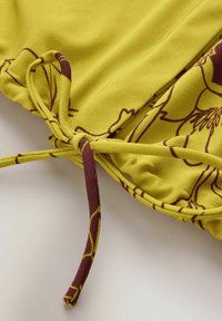 Żółta tkanina o subtelnym połysku, zdobiona kwiatowym wzorem w kolorze burgunda. Marszczony detal z wiązanym akcentem, podkreślający miękkość i układ materiału.