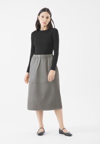 Schwarzes langärmeliges Oberteil, kombiniert mit einem grauen Leder-Midirock mit elastischem Bund. Das Modell trägt schwarze Ballerinas. Einfaches, modernes Design.