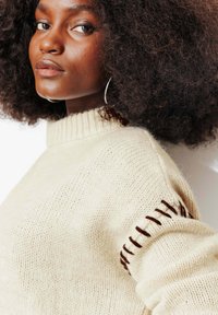 Pull en maille beige avec un col montant côtelé, orné de coutures décoratives marron sur les épaules. Texture douce, coupe décontractée.