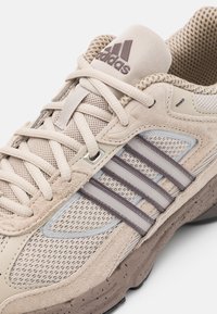 Beige atletiska skor med mesh och mocka material. Har tre bruna ränder, en texturerad sula och en framträdande vadderad plös.
