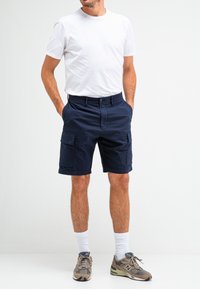Marineblauwe cargoshorts met twee zijzakken, vlakke voorkant en omgeslagen manchetten, gecombineerd met een witte T-shirt met ronde hals en grijze sneakers.