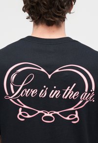 T-shirt preta de algodão com um grande coração cor-de-rosa e o texto "O amor está no ar" em fonte decorativa.