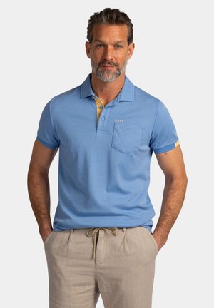 Man met baard draagt lichtblauw gestreept poloshirt en beige broek met koord, staat met handen in zakken tegen een effen achtergrond.