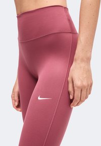 Άτομο που φοράει αθλητικό κολάν Nike με ψηλή μέση σε μοβ χρώμα και λευκό σήμα swoosh στον μηρό, με το αριστερό χέρι να ακουμπάει στον γοφό.
