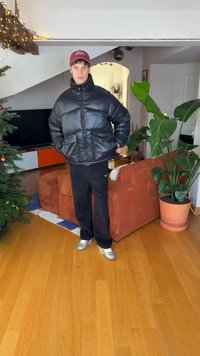 Fekete pehelykabát magas gallérral, cipzáras záródással és ráncolt textúrával. Fekete nadrággal és ezüst színű tornacipőkkel viselve egy nappali környezetben.