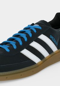 Baskets en cuir noir avec des bandes blanches, des lacets bleus, le texte "SPEZIAL" sur le côté et une semelle en gomme translucide.