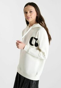Witte hoodie met een zwart logo op de voorkant, voorzien van een trekkoord en geribbelde zoom. Het model draagt het met een gestructureerde zwarte rok.