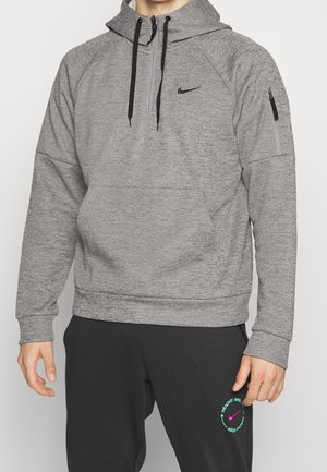 Mand iført en grå Nike hættetrøje med sorte snore og sorte Nike joggingbukser med et rundt logo på venstre lår.