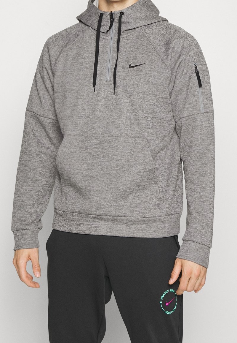 Homme portant un sweat à capuche gris Nike avec des cordons noirs et un pantalon de survêtement noir Nike avec un logo circulaire sur la cuisse gauche.