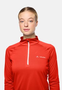 Junge Frau mit zurückgepulltem blonden Haar, trägt ein rotes Vaude-Sportoberteil mit Reißverschluss, schaut geradeaus mit einem neutralen Ausdruck.