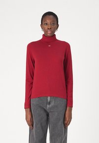 Maglione a collo alto rosso con texture a coste e un piccolo logo, abbinato a jeans grigi dal fit morbido. Design semplice, maniche lunghe.