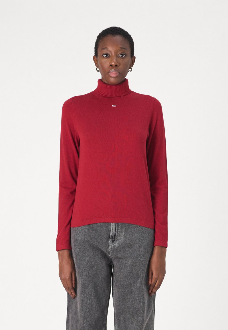 Maglione a collo alto rosso con texture a coste e un piccolo logo, abbinato a jeans grigi dal fit morbido. Design semplice, maniche lunghe.