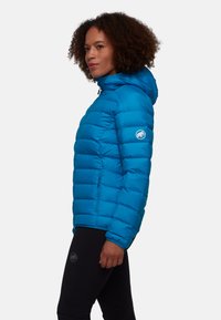 Veste en duvet bleue avec capuche, présentant des sections matelassées, des poches zippées à l'avant et un logo sur la manche. Matériau léger et isolant.