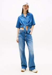 Camicia di denim cropped con nodo frontale, abbinata a jeans a gamba larga blu. Un cappellino bianco e sneaker a fantasia completano l'outfit casual.