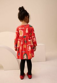 Rode jurk met lange mouwen en feestelijke patronen, waaronder sneeuwmannen en peperkoek, gecombineerd met zwarte maillots en rode glinsterende schoenen.