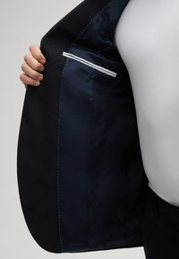 Zwarte blazer met een marineblauwe satijnen voering. Heeft witte accenten op de zak en een subtiel patroon op de voering. Soepele textuur.