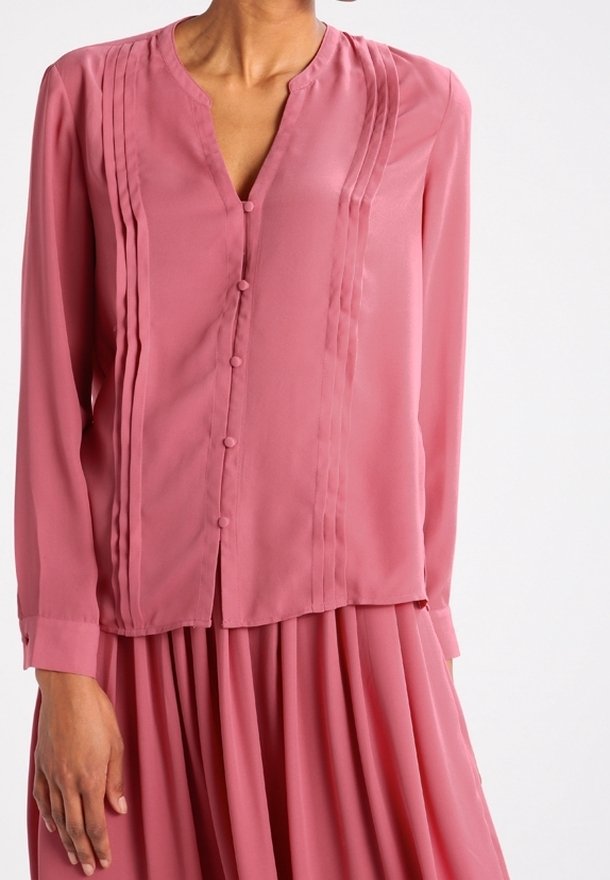 Woman wearing a long-sleeve dusty pink blouse with vertical pleats and fabric-covered buttons, paired with a matching pleated skirt.