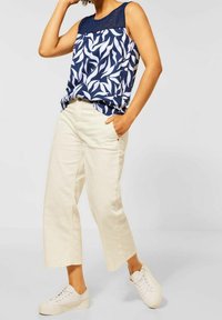 Navyblå ärmlös topp med vitt bladmönster och spetsdetaljer vid halsringningen, tillsammans med ljusbeiga vidbyxor och vita sneakers.