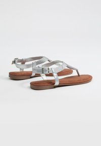 Sandalias planas con correas metálicas plateadas y una plantilla de ante marrón. Cuenta con un cierre de hebilla y un diseño minimalista.