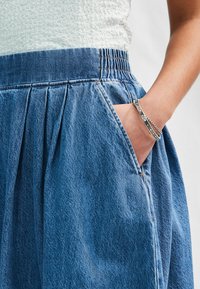 Fustă din denim cu un talie elastică, pliseuri și buzunare laterale. Culoare albastru deschis cu o textură distinctă. Brățară argintie la încheietură.