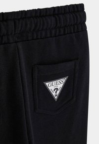 Schwarze Sweatpants mit geripptem Bündchen und einem dreieckigen Patch mit dem "GUESS"-Logo auf der hinteren Tasche. Weiche Stoffstruktur sichtbar.