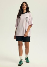 Przesadna różowa siateczkowa koszulka z czarnymi akcentami i grafiką, zestawiona z czarnymi szortami i zielonymi trampkami, w komplecie z białymi skarpetami w paski.