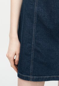 Αριστερό χέρι που ακουμπά δίπλα σε σκουρό μπλε τζιν φούστα με ορατές κάθετες και οριζόντιες ραφές.