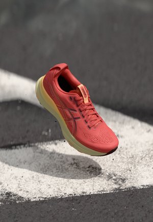 Chaussure de sport rouge avec une tige en maille, semelle intermédiaire jaune et surface texturée ; présente des lacets et des accents de logo. Positionnée au-dessus d'une surface noire.