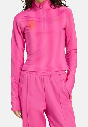 Femme portant un haut zippé manches longues rose vif et un pantalon assorti avec des poches, tous deux avec des logos Adidas subtils.