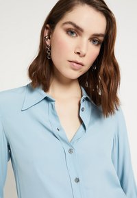 Mladá žena s hnědými vlasy po ramena má na sobě světle modrou košili na knoflíky a stříbrné náušnice na pozadí v jednobarevné, světlé barvě.