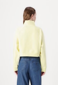 Abgeschnittenes, gelbes Sweatshirt mit hohem Kragen und gerippten Bündchen, kombiniert mit hochgeschnittenen blauen Jeans mit Gesäßtaschen.