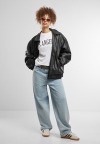 Blouson en cuir noir sur un t-shirt graphique blanc, associé à un jean large bleu clair et des baskets beiges à rayures foncées.