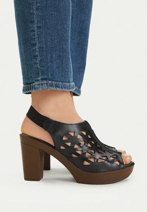 Černá kožená sandála na podpatku se sponou na patě a geometrickými výřezy, nošená s modrými džínami, které odhalují kotníky a prsty na nohou.