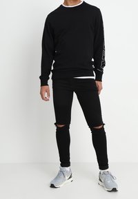 Sudadera negra con tejido texturizado, cuello redondo y detalle gráfico en la manga, combinada con jeans negros ajustados desgastados y zapatillas claras.