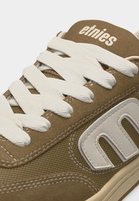 Καφέ αθλητικό παπούτσι Etnies με λευκά κορδόνια, λεπτομέρειες από δίχτυ και σουέτ, και μεγάλο λευκό λογότυπο "E" στη πλευρά.